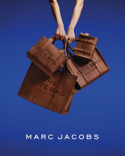 Una donna posa all'aperto con in mano una borsa marrone Marc Jacobs, si appoggia all'indietro con la testa inclinata e indossa una giacca color castagna e jeans blu. Nell'immagine compaiono le scritte "THE TOTE BAG MARC JACOBS" e "MARC JACOBS".