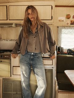 Una donna con lunghi capelli biondi ondulati indossa un mantello con cappuccio marrone e una cintura nera, si trova di fronte a una parete di legno rustica e guarda con sicurezza la fotocamera.