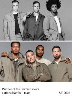 Un uomo con capelli scuri e ricci, che indossa un blazer nero sopra un maglione verde a coste, sta in piedi con le braccia conserte davanti a uno sfondo grigio uniforme e guarda dritto davanti a sé con un'espressione neutra.