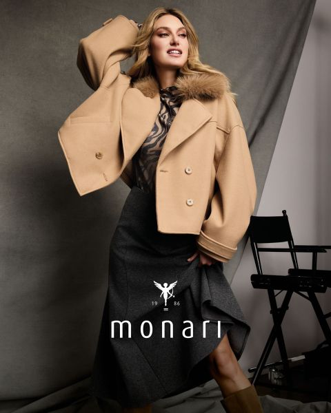 Una donna con un mantello di pelliccia sintetica sopra un top leopardato e una gonna marrone è seduta e in posa; sotto sono visibili "monari" e il logo.