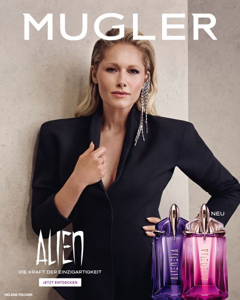 Una donna con una pelle scura e radiosa e lisci capelli neri posa davanti a uno sfondo viola. Davanti a lei ci sono due flaconi di profumo Alien di Mugler. Il testo recita "MUGLER" e "ALIEN EXTRA INTENSE LE FRAGRANZE RICARICABILI".