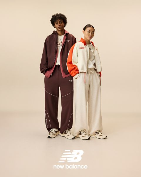 Un uomo e una donna posano davanti a insegne al neon. L'uomo indossa una tuta beige, la donna una salopette di jeans. Entrambi indossano scarpe da ginnastica New Balance. La scena è illuminata da diverse luci al neon blu e rosse.