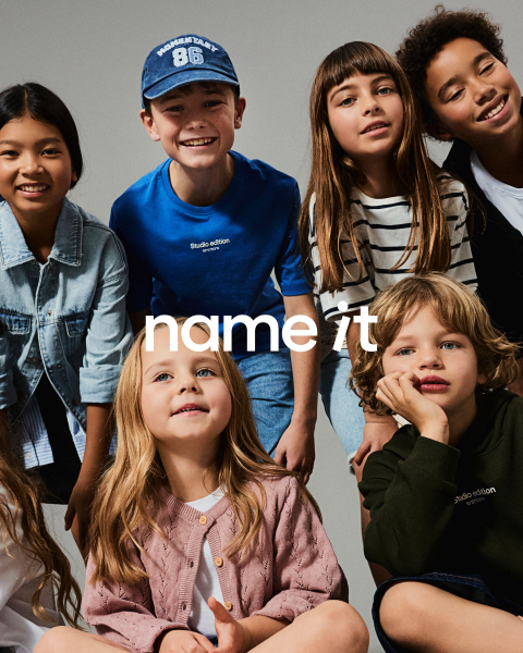 Un gruppo di otto bambini diversi è in piedi insieme su uno sfondo grigio, tutti indossano abiti casual coordinati in colori neutri e pastello. Il testo "name it" appare al centro dell'immagine.