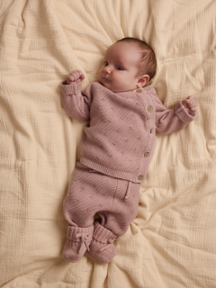 Un neonato è sdraiato su una coperta bianca e indossa un maglioncino di lana rosa chiaro con colletto alla Peter Pan e pantaloni abbinati, oltre a calzini color crema.