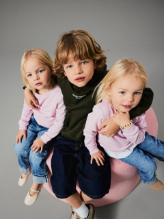 Due bambini piccoli stanno in piedi tenendosi per mano davanti a uno sfondo marrone uniforme. Un bambino indossa una giacca rosa e una gonna, l'altro jeans blu e una camicia blu con la scritta "L'AMORE È BAMBINO". Entrambi guardano in su con curiosità.