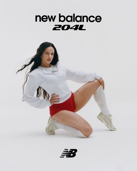 Un uomo e una donna posano davanti a insegne al neon. L'uomo indossa una tuta beige, la donna una salopette di jeans. Entrambi indossano scarpe da ginnastica New Balance. La scena è illuminata da diverse luci al neon blu e rosse.