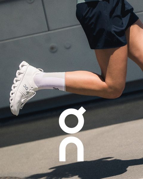 Due sneaker del marchio "On" sono presentate su un ramo strutturato di fronte a un cielo colorato al tramonto. Una scarpa è nera con accenti grigi e l'altra è bianca con dettagli più scuri. Sotto è visibile il logo "On".