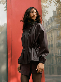 Una donna in un completo marrone oversize con cintura nera e gioielli d'oro cammina con sicurezza attraverso una strada cittadina, portando una grande borsa nera. Sullo sfondo si vedono persone sedute ai tavolini di un caffè.