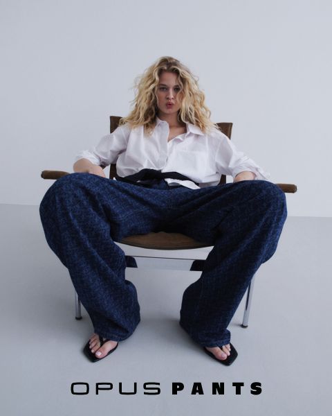 Persona in camicia bianca leggera e jeans, mani sui fianchi. In basso c'è il testo "Opus Pants". I capelli sono leggermente scompigliati dal vento, sfondo bianco.