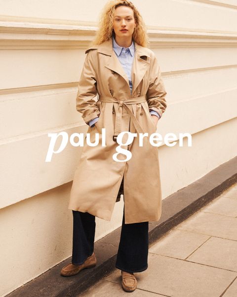 Una persona con lunghi capelli rossi, occhiali da sole, un top verde chiaro, pantaloni beige e scarpe da ginnastica blu è seduta su un terreno roccioso sotto un cielo azzurro. Le parole "Paul Green" sono sovrapposte all'immagine.