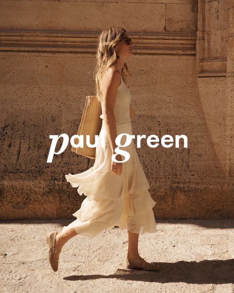 Una persona con lunghi capelli rossi, occhiali da sole, un top verde chiaro, pantaloni beige e scarpe da ginnastica blu è seduta su un terreno roccioso sotto un cielo azzurro. Le parole "Paul Green" sono sovrapposte all'immagine.