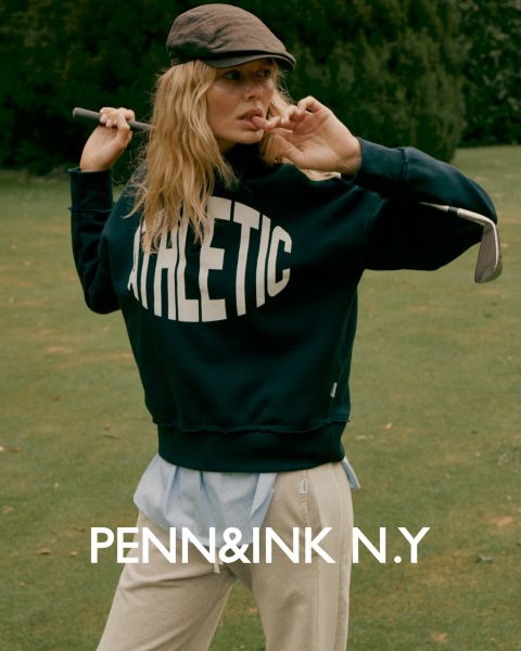 Una donna con lunghi capelli ondulati indossa un abito scuro e sandali con tacco alto, e posa con sicurezza in uno studio con sfondo monocolore. Il testo in basso recita "PENN&INK N.Y."