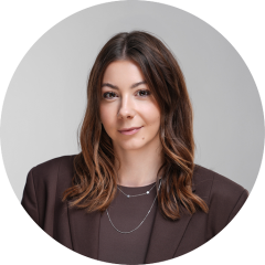 Donna con lunghi capelli castani, che indossa un blazer marrone e collane a strati, sorride dolcemente davanti a uno sfondo grigio neutro.