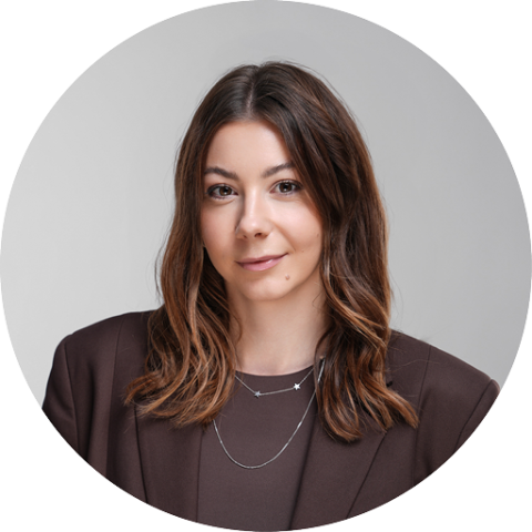 Donna con lunghi capelli castani, che indossa un blazer marrone e collane a strati, sorride dolcemente davanti a uno sfondo grigio neutro.