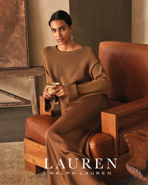 Una donna con lunghi capelli ondulati è appoggiata su un divano bianco, indossa un top rosso brillante e pantaloni abbinati. La calda luce del sole proietta ombre sulla scena. In basso si legge "LAUREN RALPH LAUREN".