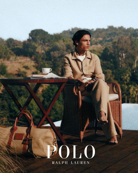 Un uomo e una donna indossano eleganti trench Ralph Lauren. L'uomo abbina il suo cappotto a un maglione colorato e pantaloni grigi, mentre la donna indossa una camicia a righe, guanti marroni e una cintura larga. "POLO RALPH LAUREN" è mostrato in caratteri bianchi.
