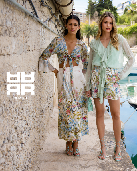 Due donne posano con stile in un teatro con sedili rossi. Una è seduta sullo schienale di un sedile e indossa un abito fantasia, un cappotto nero e stivali. L'altra è in piedi accanto a lei con un maglione blu navy e una gonna nera. Sopra di loro appare il logo "RIANI".