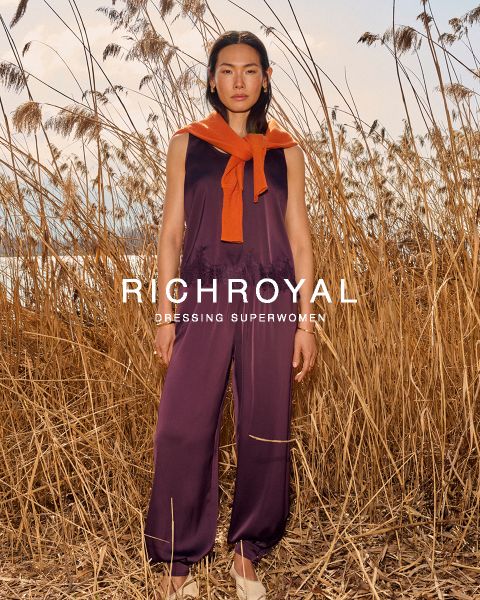 Una donna in camicia azzurra e pantaloni grigi si tiene una palla di neve davanti a un occhio. Posa in una stanza con pareti in legno e un poster di sci, e indossa grandi orecchini. Il testo recita "RICHROYAL DRESSING SUPERWOMEN".