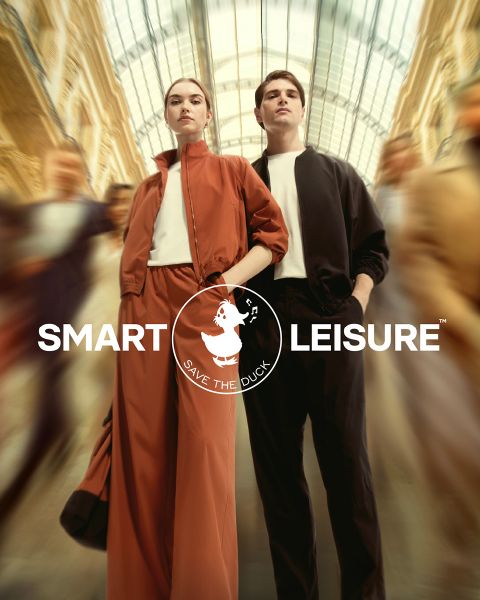 Una modella cammina su un sentiero coperto di foglie. Indossa una giacca di pelliccia arancione, una gonna abbinata e un cappello con calze arancioni e calzini color pesca. Nell'angolo si vede il logo di "Save the Duck".