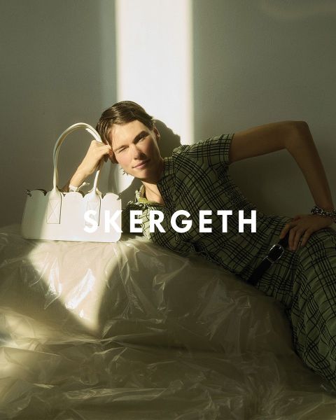 Persona in un blazer nero sopra una camicia a righe e jeans blu, che tiene una borsa nera. Il testo "JULIA SKERGETH" è chiaramente visibile sovrapposto all'immagine.