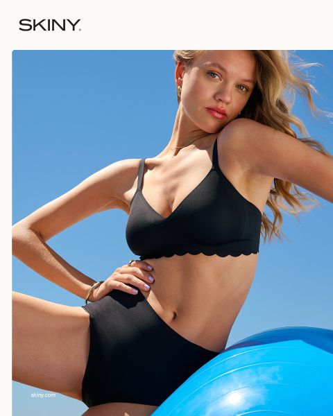 Una donna con i capelli intrecciati allaccia un reggiseno rosa chiaro con chiusura frontale su uno sfondo rosso scuro. La parola "SKINY" appare nella parte inferiore dell'immagine.