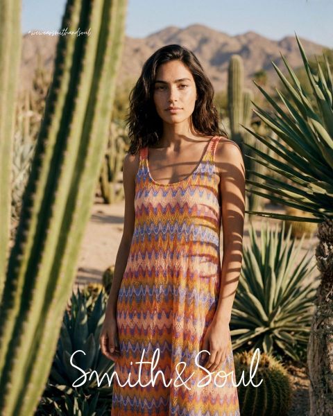 Una donna con una camicia rosa e un cardigan con pantaloni bianchi posa davanti a uno sfondo bianco. Il testo recita "Smith & Soul" in basso.