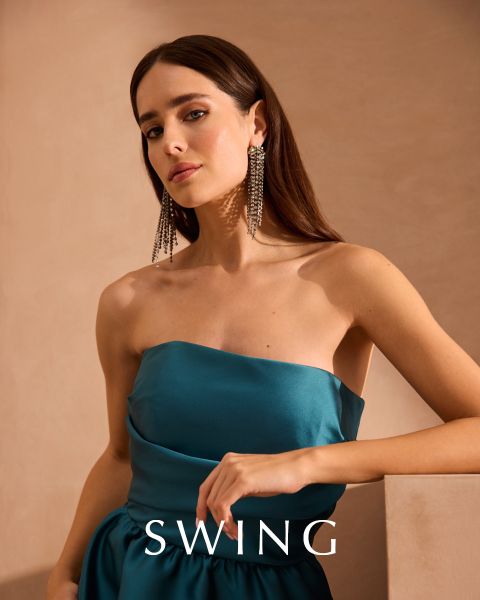 Una donna in un abito di seta color verde salvia è seduta su una sedia color crema davanti a uno sfondo floreale. Indossa tacchi dorati, orecchini vistosi e un braccialetto. La parola "SWING" appare nella parte inferiore dell'immagine.