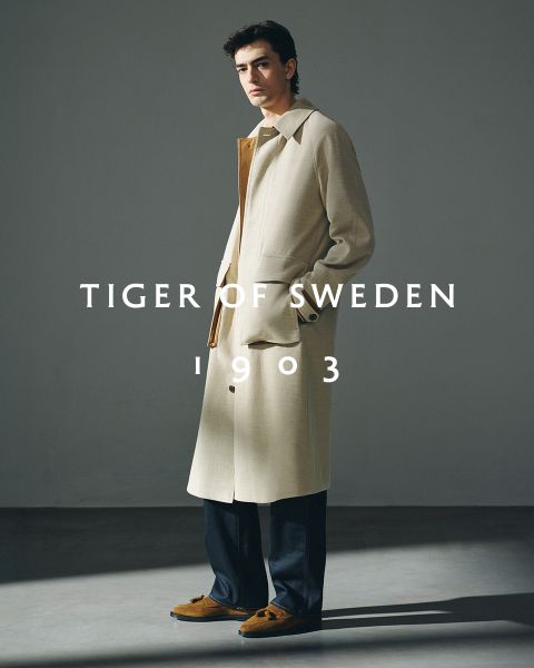 Due uomini in cappotti eleganti sono in piedi all'aperto vicino all'acqua sotto un cielo limpido. Il testo "TIGER OF SWEDEN 1903" è centrato nell'immagine.