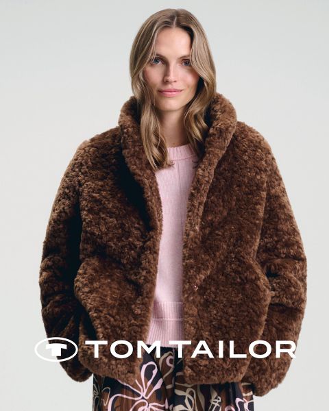 Una donna sorridente con un maglione color castagna e un uomo con un maglione blu navy sono in piedi fianco a fianco su una spiaggia. Entrambi indossano jeans. Il cielo è limpido e blu. Il logo "Tom Tailor" appare in basso.