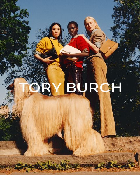 Una donna con un top scintillante e una gonna verde è seduta accanto a scatole regalo verdi e tiene in mano una borsa fiorita di Tory Burch; dietro di lei c'è un albero di Natale decorato.