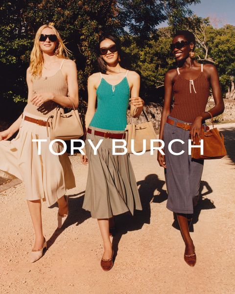 Una donna con un top scintillante e una gonna verde è seduta accanto a scatole regalo verdi e tiene in mano una borsa fiorita di Tory Burch; dietro di lei c'è un albero di Natale decorato.