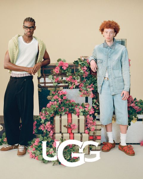 Un paio di stivali UGG in pelle scamosciata marrone con interno in peluche appoggiati su una superficie grigia. Il logo UGG è ben visibile nella parte inferiore dell'immagine.