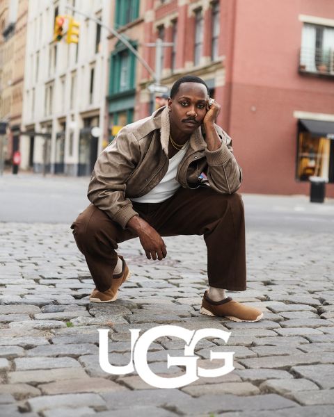 Un paio di stivali UGG in pelle scamosciata marrone con interno in peluche appoggiati su una superficie grigia. Il logo UGG è ben visibile nella parte inferiore dell'immagine.