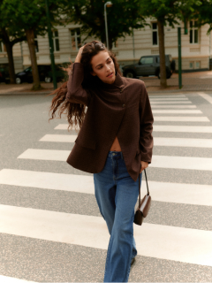 Una donna con i capelli ricci è in piedi accanto a una parete piastrellata. Indossa un cardigan grigio chiaro e soffice, pantaloncini di pelle neri a vita alta e collant neri, con le mani in tasca e un'espressione sicura di sé.