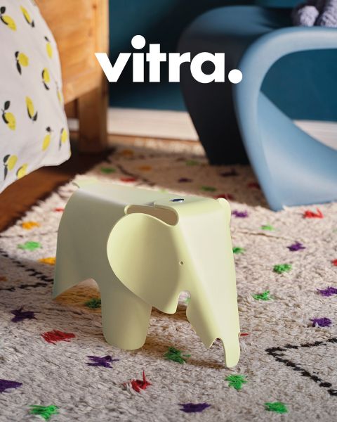 Un soggiorno moderno presenta un divano a righe rosa-beige, una sedia bianca con gambe in legno e un tavolino da caffè rotondo in legno. Sul tavolo c'è un vaso con fiori colorati, sopra c'è scritto "vitra." sulla parete. Il pavimento è ricoperto da un morbido tappeto.