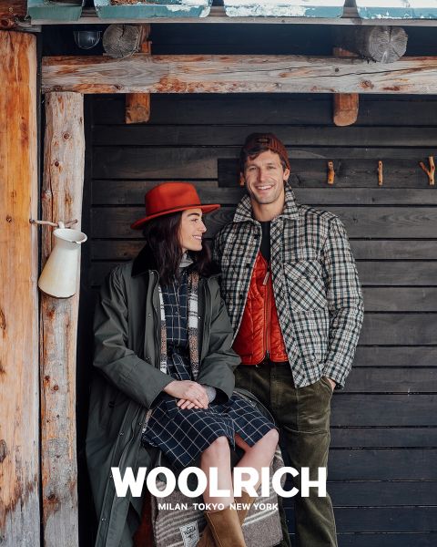 Una persona che indossa un cappotto invernale marrone chiaro, un berretto di lana blu e una camicia a quadri blu sopra una felpa con cappuccio, si trova di fronte a uno sfondo a tinta unita. Sotto appare la parola "Woolrich" insieme alle città di Milano, Tokyo e New York.