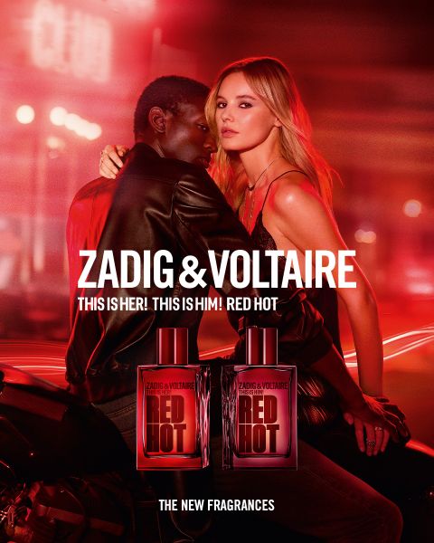 Donna con lunghi capelli rossi e abito nero seduta su un letto in una stanza con una gabbia per uccelli e una grande finestra. Testo: "Zadig & Voltaire" e "Zadig: La nuova fragranza ricaricabile". Logo con ali e un ferro di cavallo.