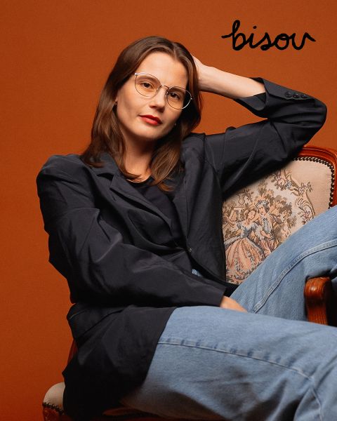 Una donna con occhiali, blazer scuro e jeans è seduta su una sedia decorata davanti a uno sfondo color ruggine, sopra di lei la parola "Bisou" è scritta in nero.
