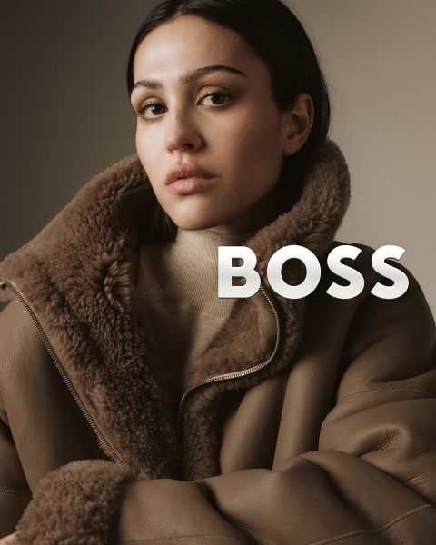 Una donna con un cappotto shearling marrone guarda con sicurezza la fotocamera su uno sfondo neutro. La parola "BOSS" è sovrapposta in grassetto bianco accanto alla sua spalla.