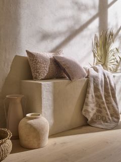 Uno spazio minimalista presenta una panca in pietra beige con due cuscini marroni e beige testurizzati, un plaid a motivi e erbe essiccate. Un cesto intrecciato, un vaso in ceramica rustica e una brocca alta adornano il pavimento sotto una luce naturale soffusa.