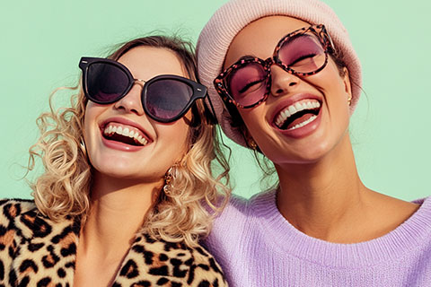 Zwei lächelnde Frauen mit modischer Sonnenbrille und Winterkleidung. Die eine hat lockiges Haar und einen Mantel mit Leopardenmuster, die andere trägt eine rosa Mütze und einen lila Pullover. Sie stehen vor einem pastellgrünen Hintergrund und wirken fröhlich und stilvoll.