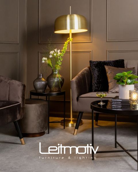 Un soggiorno elegante con una lampada da terra dorata, mobili in velluto scuro e vasi decorativi su tavolini. Su un tavolino da caffè ci sono un bicchiere e una candela. La parete rivestita in legno crea uno sfondo elegante. In basso si trova il logo "Leitmotiv Mobili & Illuminazione".