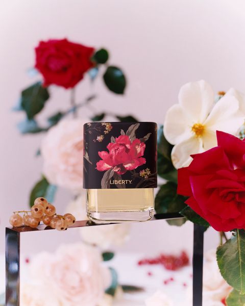 Flacone di profumo con design floreale e la scritta "Liberty" su una superficie di vetro. Circondato da rose rosse e bianche, foglie verdi, ribes rossi e uno sfondo sfocato con altri fiori.