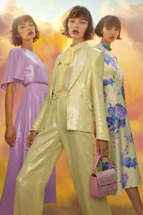 Tre modelle posano davanti a un cielo luminoso e nuvoloso. Indossano abiti color pastello: un vestito viola, un costume di paillettes giallo con papillon e un abito bianco a fiori. Una modella tiene in mano una borsetta glitterata rosa.