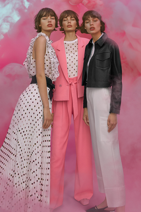 Tre donne vestite alla moda sono in piedi davanti a uno sfondo rosa e fumoso. Una indossa un vestito a pois, una un tailleur rosa con una camicia a pois e la terza una giacca nera e pantaloni bianchi. Posano con sicurezza ed emanano un'aria moderna e chic.