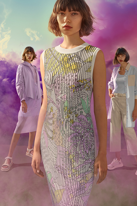 Tre donne posano davanti a uno sfondo luminoso di nuvole viola e rosa. La figura centrale indossa un abito a motivi, mentre le altre indossano abiti casual e chiari. La scena è colorata e artistica.