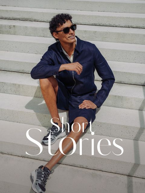 Un uomo con occhiali da sole, giacca blu navy e pantaloncini è seduto sorridendo su una scalinata esterna. Le parole "Short. Stories" appaiono in basso.