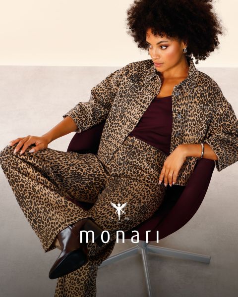 Una donna sorridente che indossa un cappotto beige, una camicia a righe, jeans blu e una sciarpa chiara è in piedi all'aperto di fronte a un edificio alto. Il logo Monari è visibile nella parte inferiore dell'immagine.