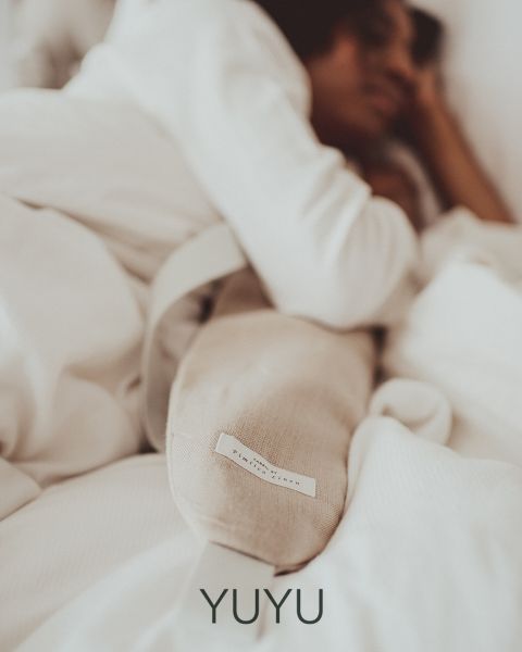 Una persona è sdraiata in un letto, avvolta in una coperta bianca, e tiene in mano un cuscino lungo e chiaro o un termoforo. L'atmosfera è tranquilla e confortevole, trasmettendo sicurezza e relax. In basso c'è la parola "YUYU".