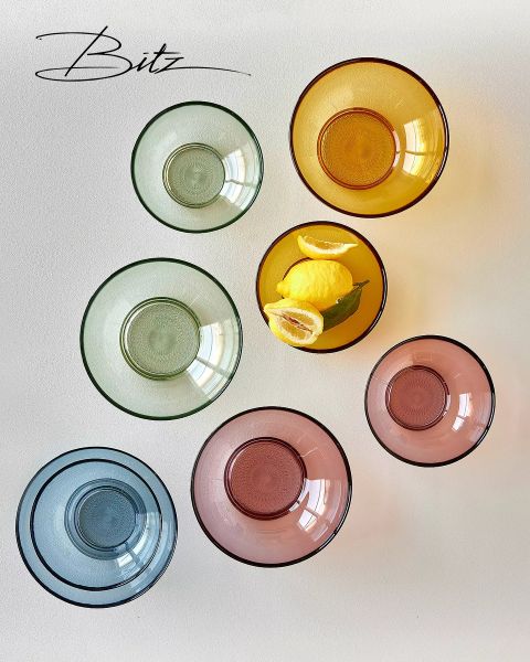 Sette ciotole di vetro colorate, disposte a cerchio su una superficie bianca. Una ciotola gialla contiene tre limoni con foglie verdi. Il logo "Bitz" è visibile nell'angolo in alto a sinistra.